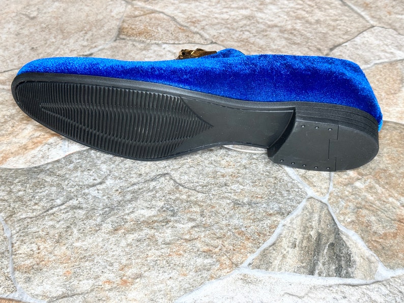 May include: A close-up of a blue velvet loafer with a black sole. The shoe features a gold-colored buckle and a textured sole. The shoe is approximately 11 inches long.