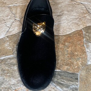 May include: Black velvet loafer with a gold-coloured panther head ornament. The shoe has a rounded toe and a low profile, suitable for formal or semi-formal occasions. The shoe is made of a soft, plush material.