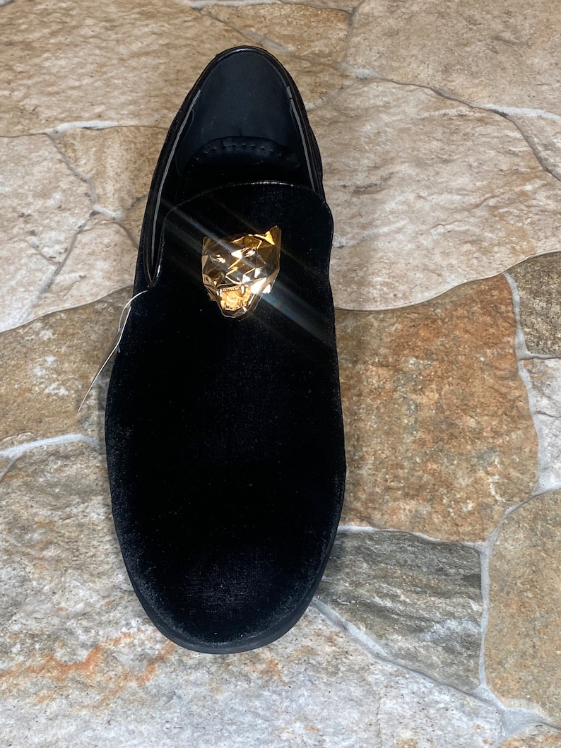 May include: Black velvet loafer with a gold-coloured panther head ornament. The shoe has a rounded toe and a low profile, suitable for formal or semi-formal occasions. The shoe is made of a soft, plush material.