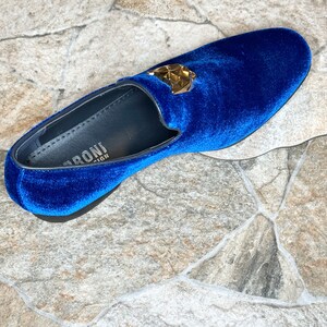 May include: A single, royal blue velvet loafer with a gold-colored geometric emblem. The shoe has a rounded toe and a black sole. The interior of the shoe is black with white text.