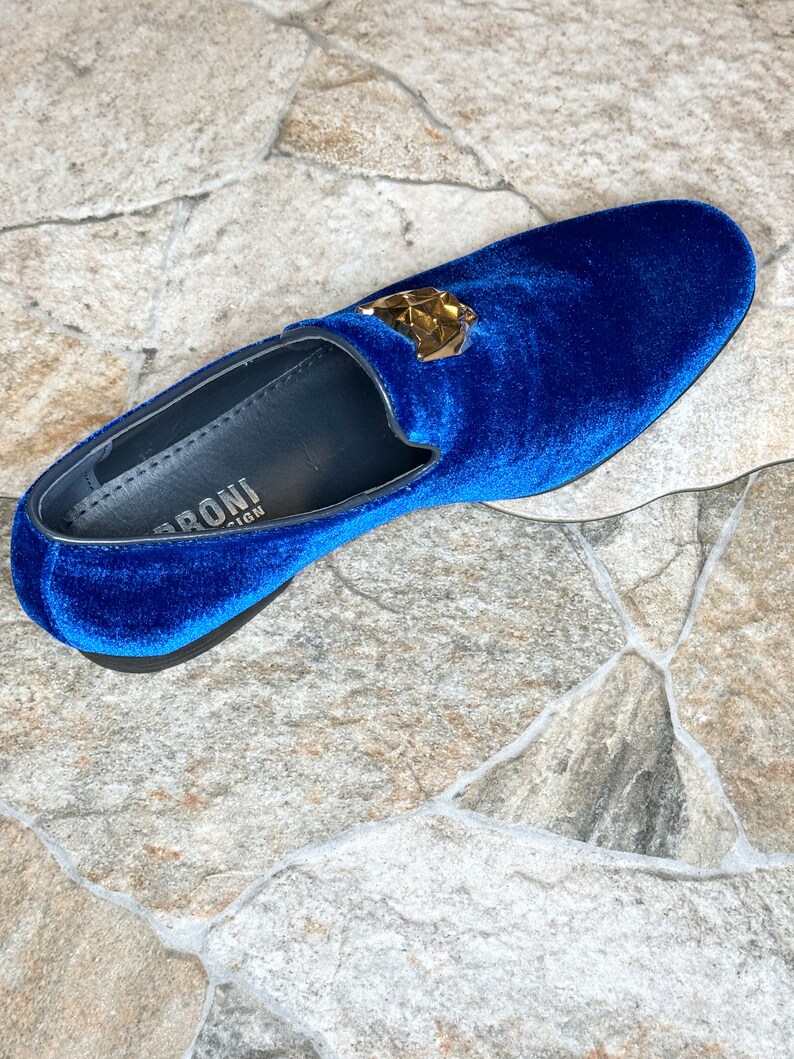 May include: A single, royal blue velvet loafer with a gold-colored geometric emblem. The shoe has a rounded toe and a black sole. The interior of the shoe is black with white text.