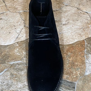 May include: A single black suede chukka boot with a lace-up closure. The boot has a rounded toe and a textured sole with white stitching. The shoe is set against a neutral stone-like surface.