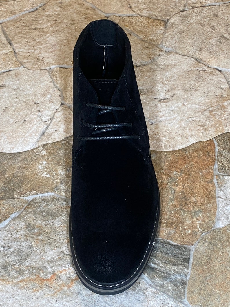 May include: A single black suede chukka boot with a lace-up closure. The boot has a rounded toe and a textured sole with white stitching. The shoe is set against a neutral stone-like surface.
