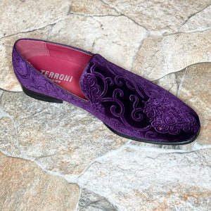 Op de afbeelding: Een enkele, paarse fluwelen loafer met een sierlijk, verhoogd bloemmotief. De schoen heeft een zwarte zool en een roze binnenkant met de tekst "TERRONI DESIGN". Geplaatst op een stenen tegelachtergrond.
