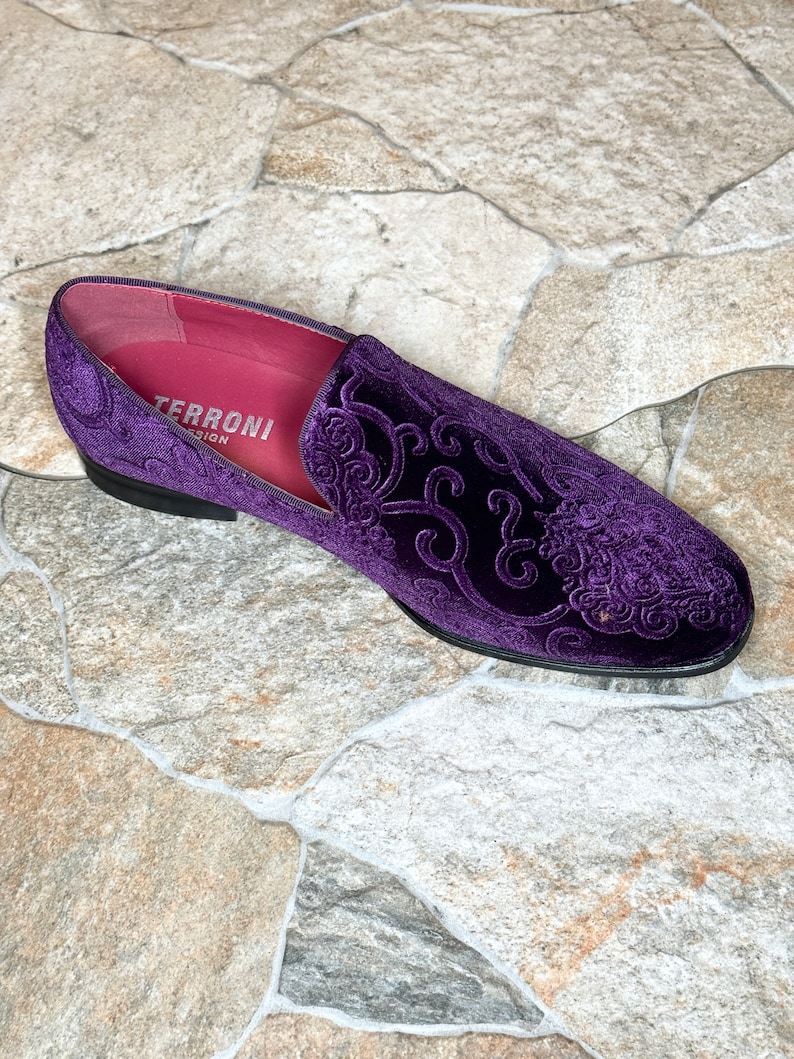 Op de afbeelding: Een enkele, paarse fluwelen loafer met een sierlijk, verhoogd bloemmotief. De schoen heeft een zwarte zool en een roze binnenkant met de tekst "TERRONI DESIGN". Geplaatst op een stenen tegelachtergrond.