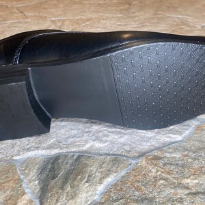 May include: A black leather dress shoe, viewed from the sole. The sole is black with a textured pattern for grip. The heel is approximately 1 inch high. The shoe is on a stone surface.