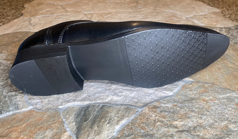 May include: A black leather dress shoe, viewed from the sole. The sole is black with a textured pattern for grip. The heel is approximately 1 inch high. The shoe is on a stone surface.