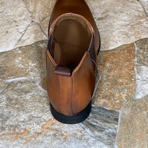 May include: A brown leather shoe with a pointed toe and a black sole. The shoe has elastic side panels and a pull tab at the back. The interior is a lighter brown color.