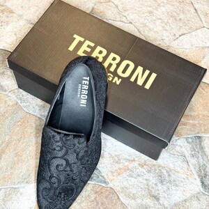 Puede incluir: Zapato negro sin cordones con un intrincado dise&ntilde;o floral, junto a una caja negra con "TERRONI DESIGN" impreso en dorado. El interior del zapato muestra el logotipo "TERRONI DESIGN". El zapato y la caja est&aacute;n sobre una superficie de piedra.