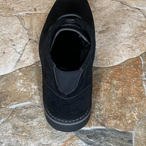 May include: Black suede ankle boot with a rounded toe and a lace-up design. The boot features a gray sole and elastic side panels for easy wear. A versatile shoe for various occasions.