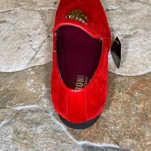 May include: A red velvet loafer with a gold-colored emblem on the vamp. The shoe has a black sole and a burgundy interior. A tag is attached to the shoe.