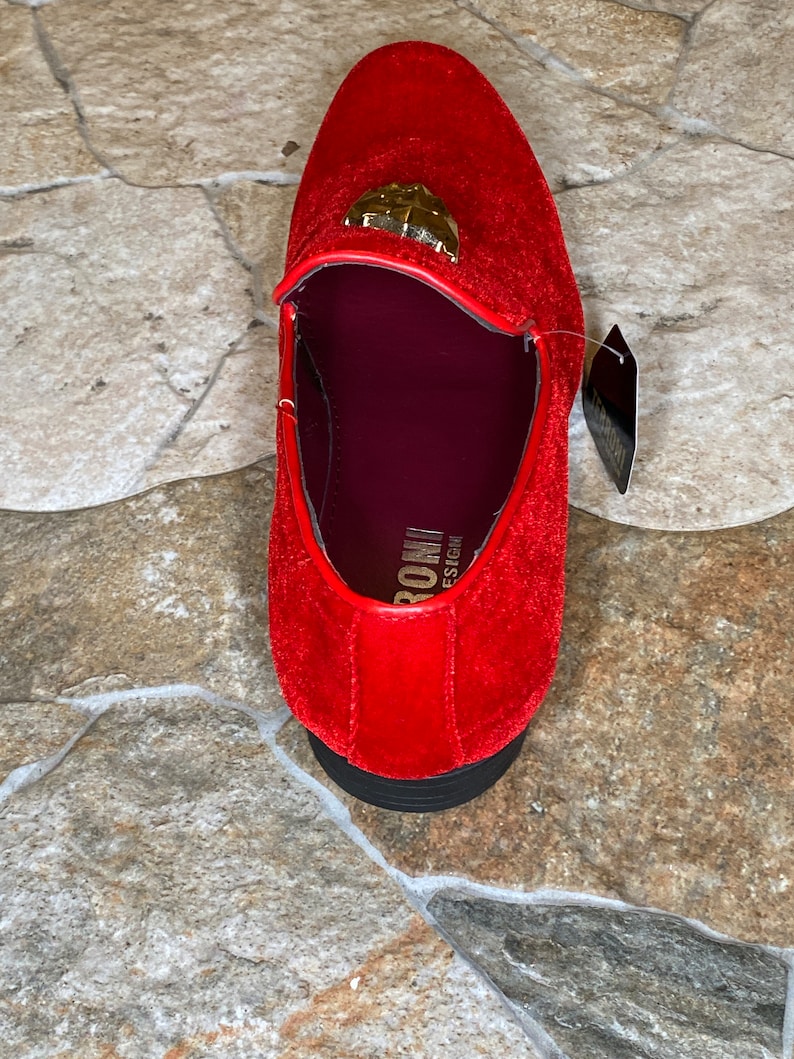 May include: A red velvet loafer with a gold-colored emblem on the vamp. The shoe has a black sole and a burgundy interior. A tag is attached to the shoe.