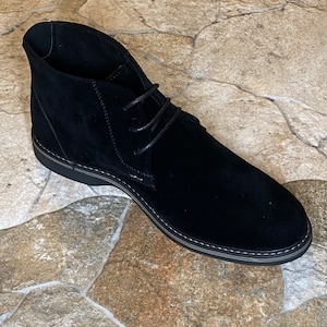May include: A black suede chukka boot with a rounded toe and a lace-up closure. The boot has a black sole with a gray trim and is set against a stone-like background.