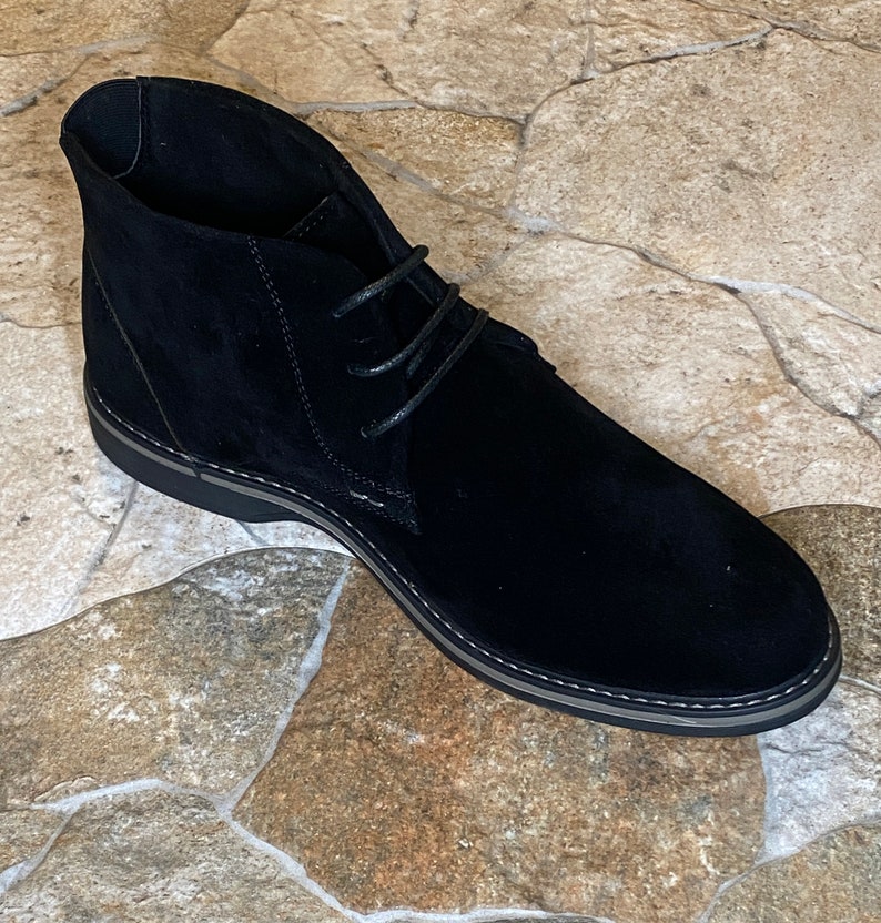 May include: A black suede chukka boot with a rounded toe and a lace-up closure. The boot has a black sole with a gray trim and is set against a stone-like background.