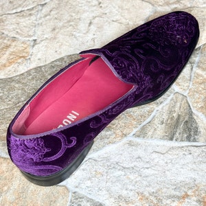 Op de afbeelding: Een enkele, dieppaarse fluwelen loafer met een sierlijk, wervelend patroon. De schoen heeft een zwarte zool en een felroze binnenkant. De schoen is een instapmodel.