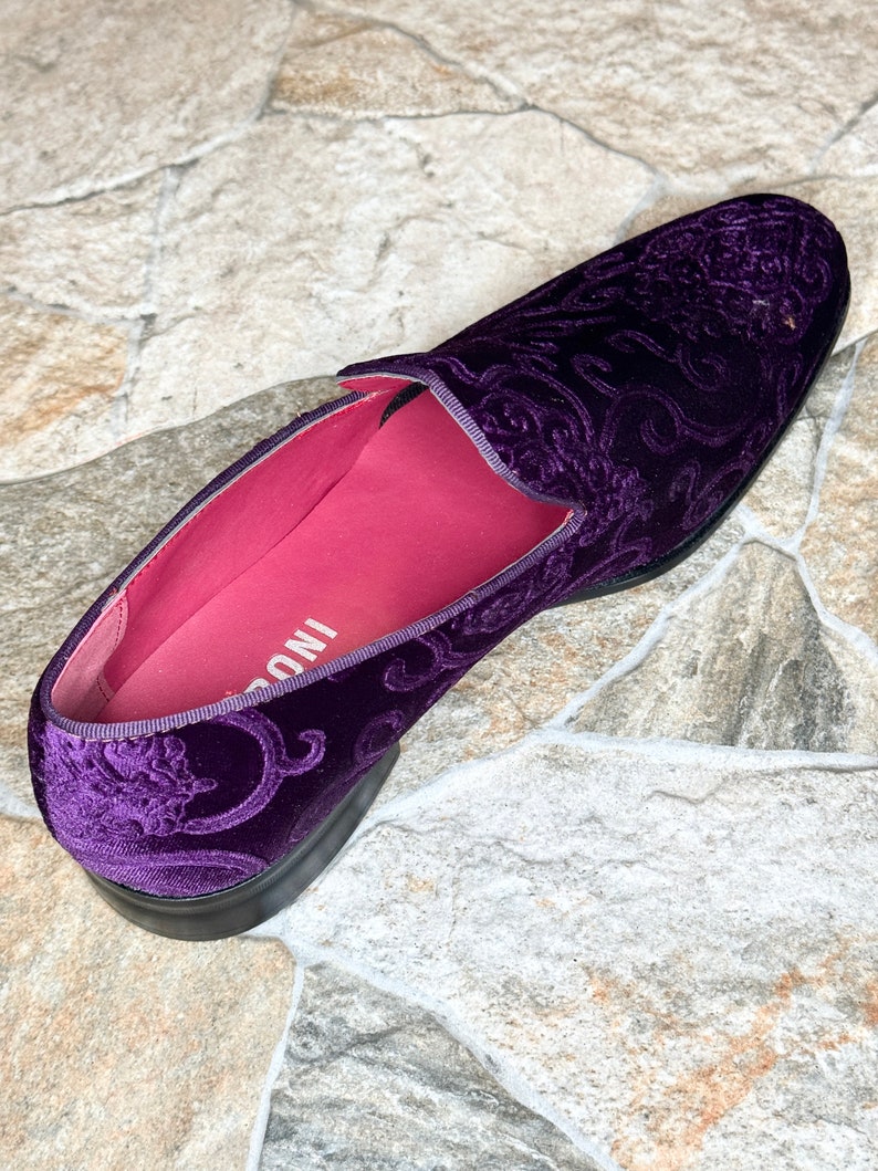 Op de afbeelding: Een enkele, dieppaarse fluwelen loafer met een sierlijk, wervelend patroon. De schoen heeft een zwarte zool en een felroze binnenkant. De schoen is een instapmodel.