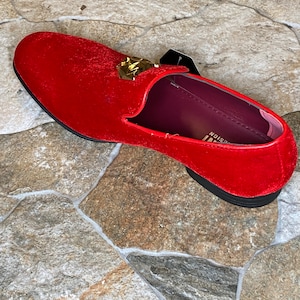 May include: A red velvet loafer with a gold-colored accent on the vamp. The shoe has a black sole and a burgundy interior. A black tag is attached to the shoe.