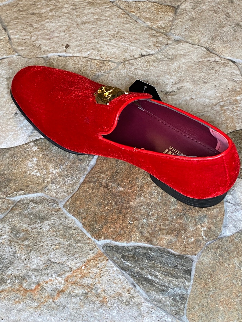 May include: A red velvet loafer with a gold-colored accent on the vamp. The shoe has a black sole and a burgundy interior. A black tag is attached to the shoe.
