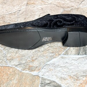 Puede incluir: Mocas&iacute;n de terciopelo negro con un patr&oacute;n ornamental y arremolinado. El zapato tiene un dise&ntilde;o elegante y de perfil bajo con una suela negra s&oacute;lida. La parte superior del zapato presenta un dise&ntilde;o floral en relieve detallado.