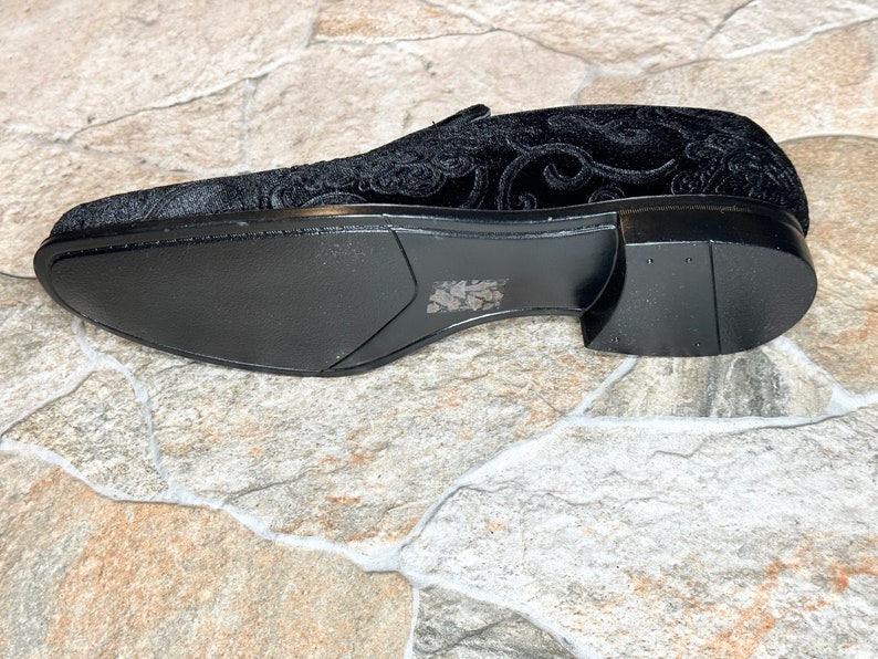 Puede incluir: Mocas&iacute;n de terciopelo negro con un patr&oacute;n ornamental y arremolinado. El zapato tiene un dise&ntilde;o elegante y de perfil bajo con una suela negra s&oacute;lida. La parte superior del zapato presenta un dise&ntilde;o floral en relieve detallado.