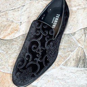 Puede incluir: Mocas&iacute;n de terciopelo negro con un elaborado dise&ntilde;o floral en relieve. El zapato presenta una puntera redondeada y un tac&oacute;n bajo. La marca "TERRONI DESIGN" es visible en el forro interior. El zapato est&aacute; sobre un fondo de baldosas de piedra.