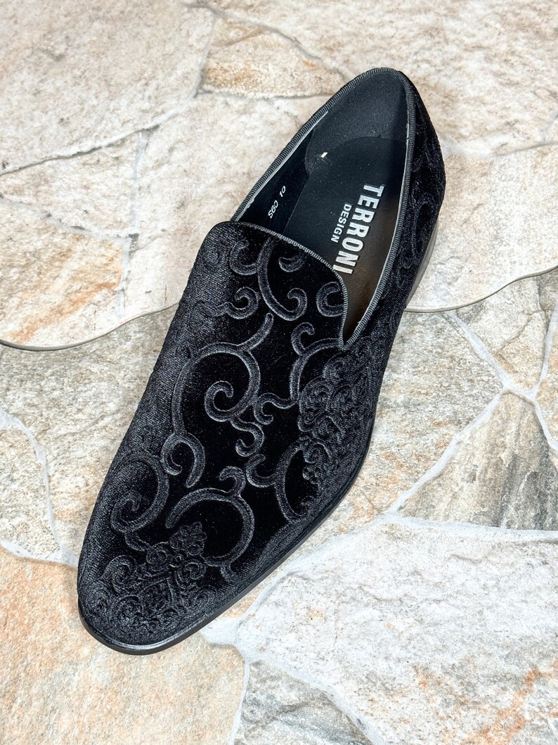Puede incluir: Mocas&iacute;n de terciopelo negro con un elaborado dise&ntilde;o floral en relieve. El zapato presenta una puntera redondeada y un tac&oacute;n bajo. La marca "TERRONI DESIGN" es visible en el forro interior. El zapato est&aacute; sobre un fondo de baldosas de piedra.