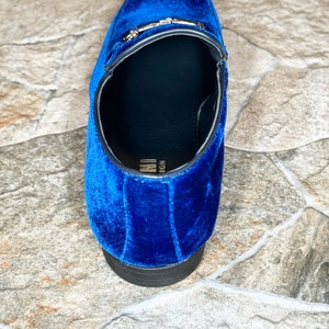 Puede incluir: Primer plano de un mocas&iacute;n de terciopelo azul. El zapato presenta una hebilla dorada y una suela negra. El mocas&iacute;n es de estilo sin cordones y el interior es negro.