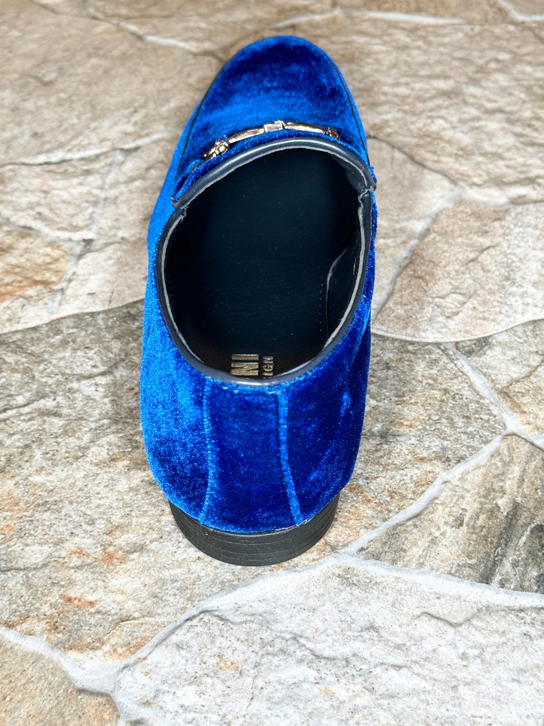 Puede incluir: Primer plano de un mocas&iacute;n de terciopelo azul. El zapato presenta una hebilla dorada y una suela negra. El mocas&iacute;n es de estilo sin cordones y el interior es negro.