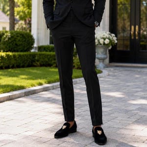 May include: A person wearing a black suit and black loafers with a gold emblem. The loafers are made of a soft, dark material. The suit is a classic, tailored style, suitable for formal occasions.
