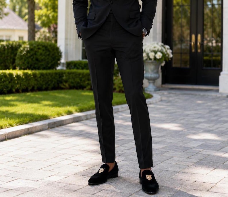 May include: A person wearing a black suit and black loafers with a gold emblem. The loafers are made of a soft, dark material. The suit is a classic, tailored style, suitable for formal occasions.