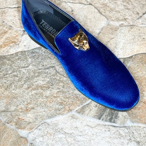 May include: A single blue velvet loafer with a gold-colored panther head emblem. The shoe has a black sole and a label that reads "TERRON ITALIAN DESIGN". The shoe is set against a stone tile background.