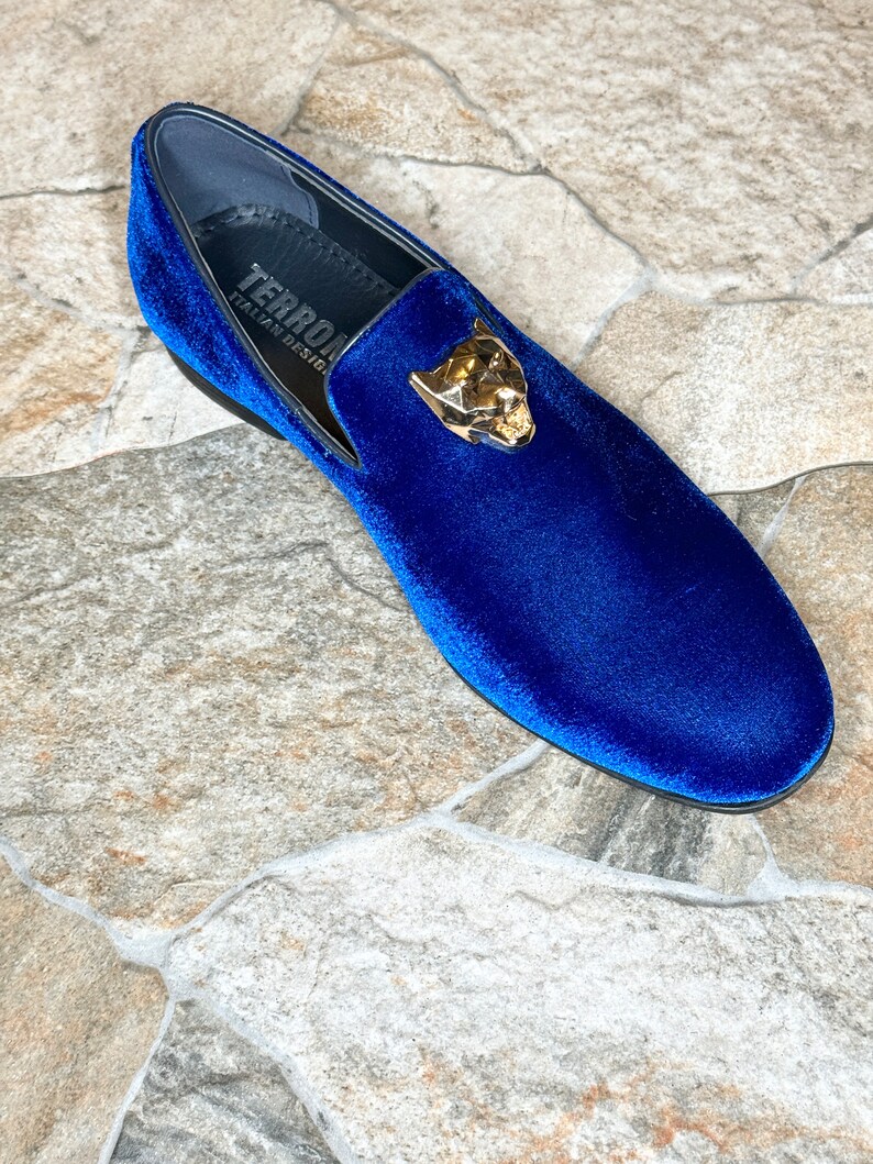 May include: A single blue velvet loafer with a gold-colored panther head emblem. The shoe has a black sole and a label that reads "TERRON ITALIAN DESIGN". The shoe is set against a stone tile background.