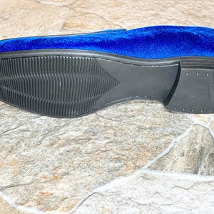 Puede incluir: Un solo mocas&iacute;n de terciopelo azul, con suela negra texturizada. El tal&oacute;n es negro liso. La suela presenta un patr&oacute;n acanalado. El zapato se encuentra sobre una superficie similar a la piedra.