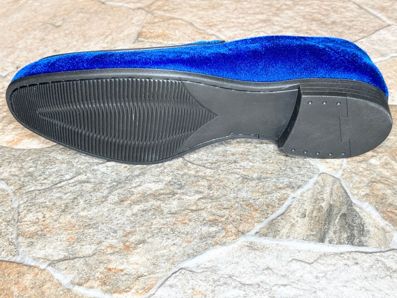 Puede incluir: Un solo mocas&iacute;n de terciopelo azul, con suela negra texturizada. El tal&oacute;n es negro liso. La suela presenta un patr&oacute;n acanalado. El zapato se encuentra sobre una superficie similar a la piedra.