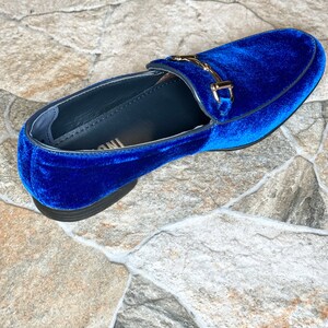 Puede incluir: Un solo mocas&iacute;n de terciopelo azul real con una hebilla dorada. El zapato tiene una punta redondeada y una suela negra. El zapato est&aacute; sobre una superficie neutra, similar a la piedra.