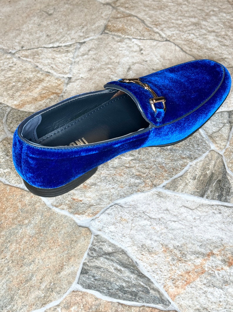 Puede incluir: Un solo mocas&iacute;n de terciopelo azul real con una hebilla dorada. El zapato tiene una punta redondeada y una suela negra. El zapato est&aacute; sobre una superficie neutra, similar a la piedra.