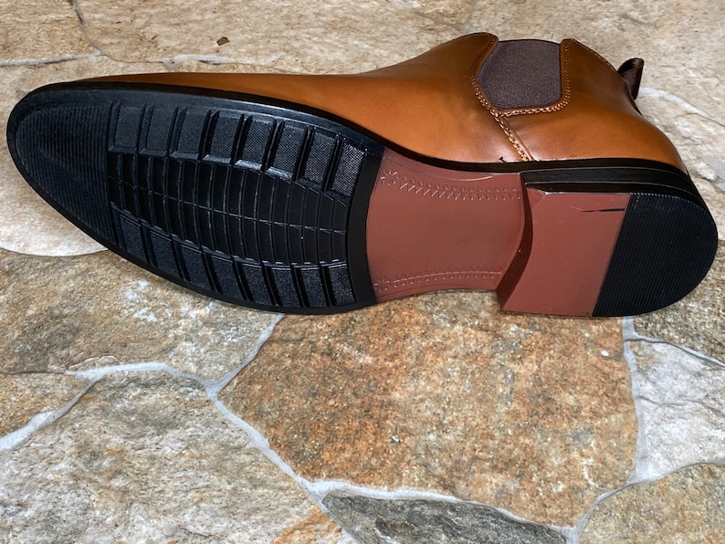 May include: A brown leather Chelsea boot with a black rubber sole. The sole has a textured pattern for grip. The boot's design includes a brown heel and a brown elastic side panel.