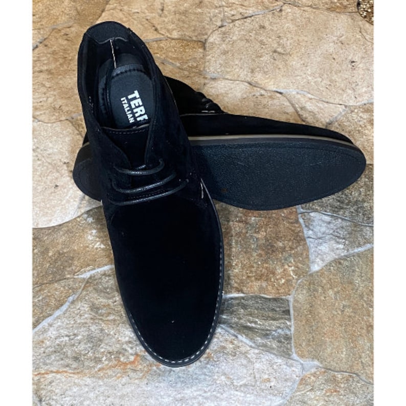 May include: A pair of black suede chukka boots with a lace-up design. The boots have a rounded toe and a stitched sole. The insole reads "TERF ITALIAN". The boots are displayed on a stone-like surface.