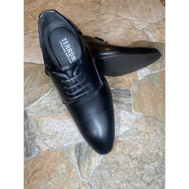 May include: Black leather dress shoes with laces. The shoes have a classic design and a smooth finish. The insole of the shoe has the text "TERRON ITALIAN DESIGN". The shoes are placed on a stone surface.
