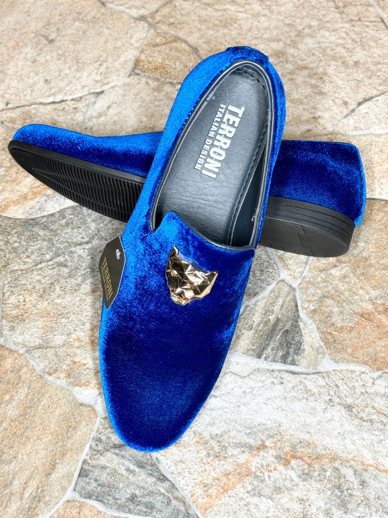 May include: Royal blue velvet loafers with a gold-colored geometric panther head detail. The shoes have a slip-on design and a black sole. The brand name "TERRONI ITALIAN DESIGN" is visible on the insole.