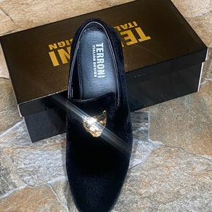 May include: Black velvet loafer with a gold-coloured panther head detail. The shoe is displayed on top of a black box with the text "TERRONI ITALIAN DESIGN". The shoe is a slip-on style.