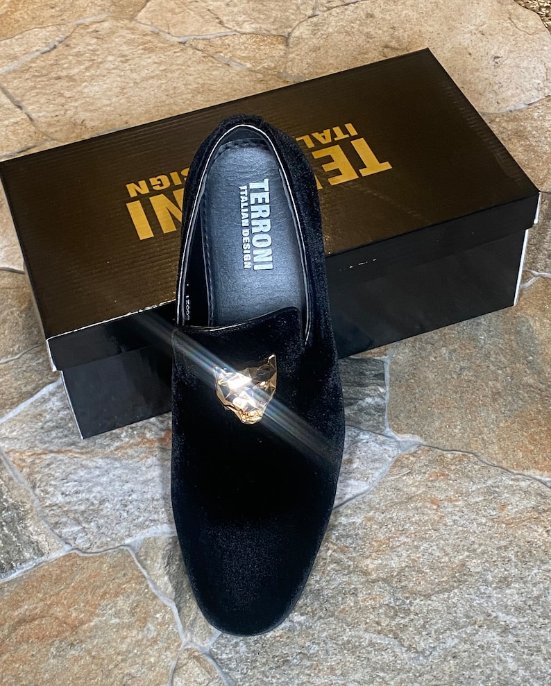 May include: Black velvet loafer with a gold-coloured panther head detail. The shoe is displayed on top of a black box with the text "TERRONI ITALIAN DESIGN". The shoe is a slip-on style.
