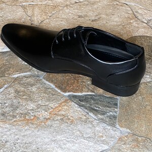 May include: A single black leather dress shoe with a pointed toe and lace-up closure. The shoe is displayed on a stone-like surface. The shoe is a classic style, suitable for formal occasions.