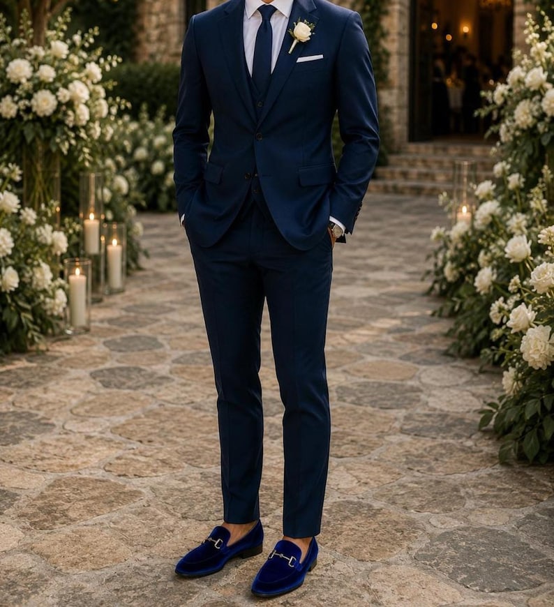 Puede incluir: Un traje de tres piezas azul marino con camisa blanca, corbata azul marino y un ojal de flores blancas. El traje se combina con mocasines de terciopelo azul marino. El fondo presenta flores blancas y velas.
