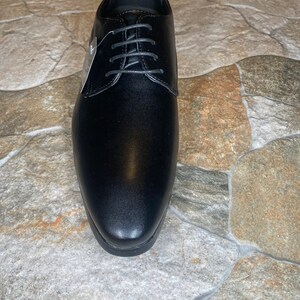 May include: A single black dress shoe with a rounded toe and a lace-up closure. The shoe has a smooth, polished finish and a small tag attached to the laces. The shoe is displayed on a stone-like surface.