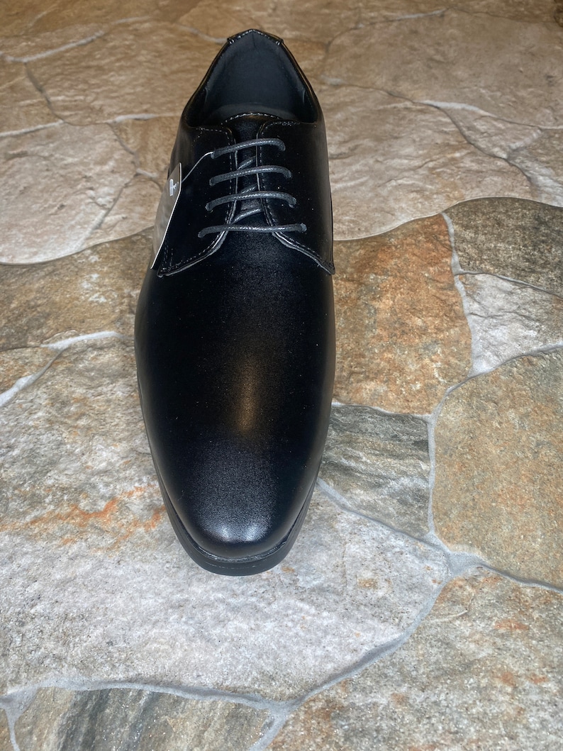 May include: A single black dress shoe with a rounded toe and a lace-up closure. The shoe has a smooth, polished finish and a small tag attached to the laces. The shoe is displayed on a stone-like surface.