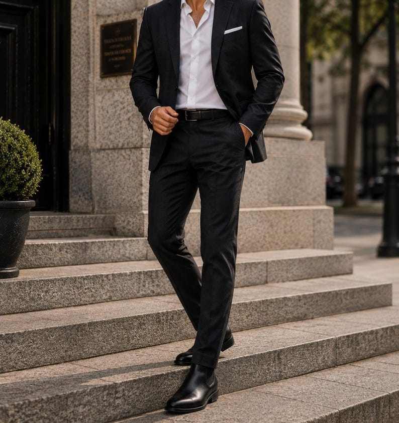 Puede incluir: Un traje gris oscuro con camisa blanca y botas de cuero negro. El traje incluye una americana, pantalones de vestir y un cintur&oacute;n negro. La persona est&aacute; de pie en escalones de piedra.