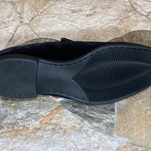 May include: Black velvet loafer shoe with a textured rubber sole. The sole has a ridged pattern for grip and a heel with a lightning bolt design. The shoe's upper is a soft, dark fabric.