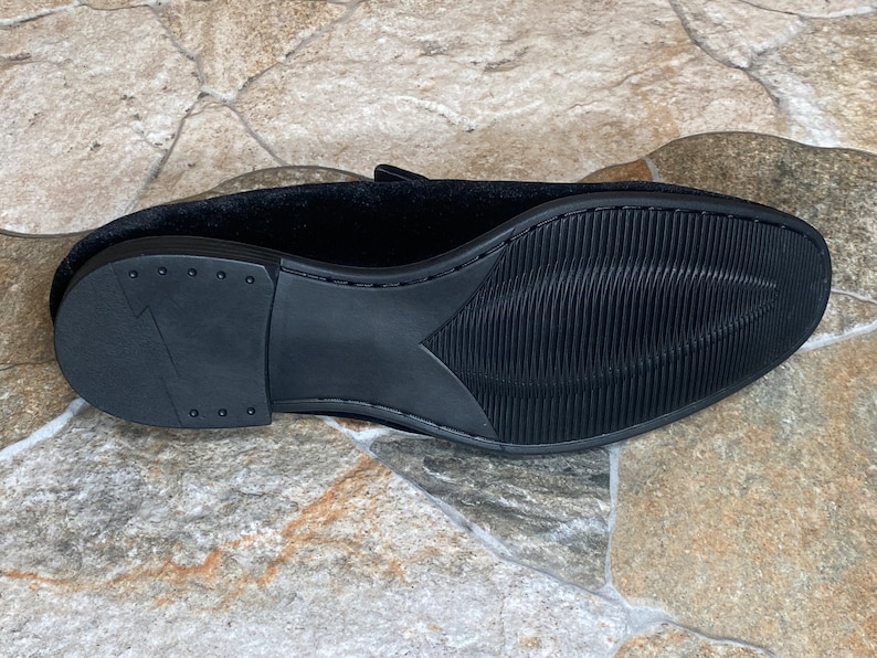 May include: Black velvet loafer shoe with a textured rubber sole. The sole has a ridged pattern for grip and a heel with a lightning bolt design. The shoe's upper is a soft, dark fabric.