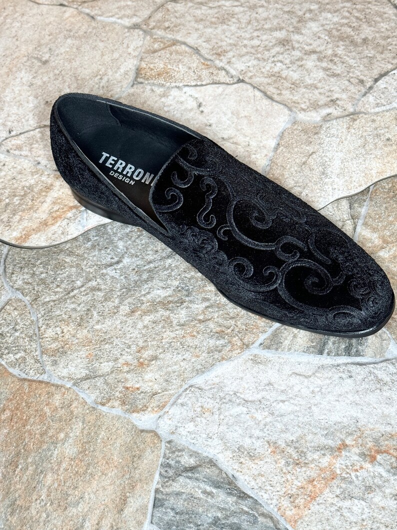 Puede incluir: Mocas&iacute;n de terciopelo negro con un dise&ntilde;o ornamental y ondulado. El zapato presenta un tac&oacute;n bajo y el texto "TERRONI DESIGN" en el interior. Un calzado elegante.